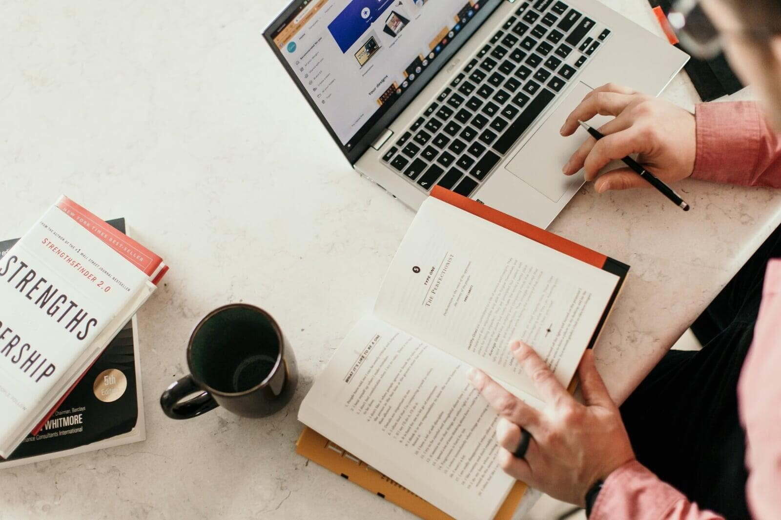 Professional reading StrengthsFinder and leadership development books while working on laptop, representing personal assessment tools used in organizational alignment and leadership coaching.